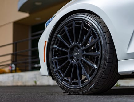 Close-up of a black Niche wheel with multiple spokes on a white car, parked on a paved surface with a blurred background of a building.