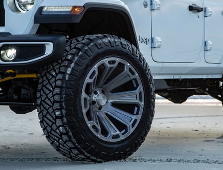 Close-up of a white Jeep with large, rugged off-road tires and stylish modern rims parked on a sandy surface.