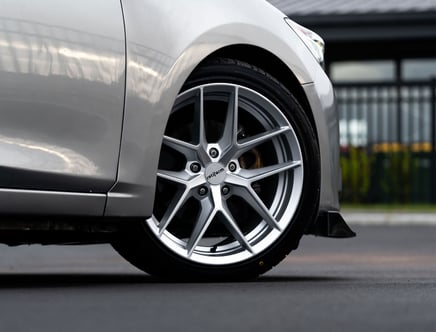 Close-up of a silver car's wheel with a shiny, multi-spoke alloy rim on a smooth road surface, part of the vehicle's body visible.