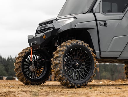 Close-up of the front section and wheels of a rugged off-road vehicle with large, muddy tires and a heavy-duty front bumper, parked on a dirt surface.