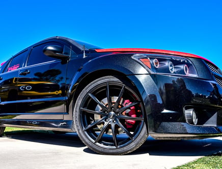 Low-angle view of a black sports car with red accents, featuring custom rims and tire, parked on a sunny day.