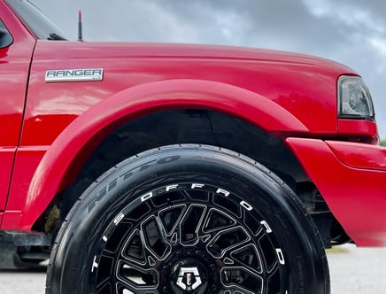 Close-up of a red Ford Ranger XLT's front wheel and fender, featuring a black rim with TIS Offroad branding and Nitto tires, against a cloudy sky background.