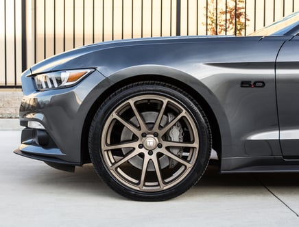 Close-up view of a gray car's front side, showcasing a sleek headlight, bronze alloy wheel, and "5.0" badge on the fender.