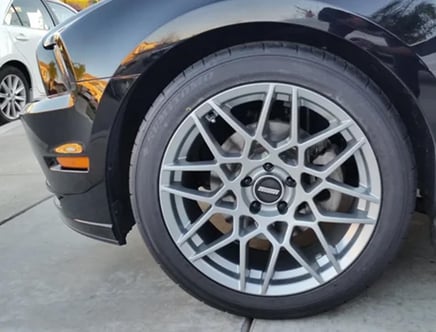 A close-up of a car's front wheel featuring a multi-spoke alloy rim design, with part of the black front bumper visible, parked on a driveway.