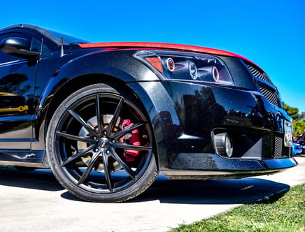 Close-up of a black sports car with a shiny exterior, showcasing its large black alloy wheel with red brake caliper, under a clear blue sky.