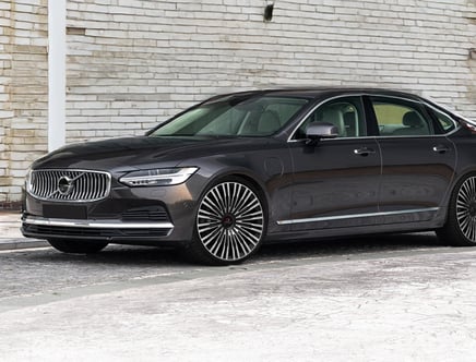 A sleek, dark-colored Volvo sedan parked in front of a white brick wall, featuring distinctive large wheels and a prominent grille, viewed from the front side angle.