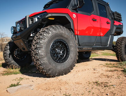 A rugged off-road vehicle with large, aggressive tread tires and a bright red and black body, parked on a dirt path under a clear blue sky.