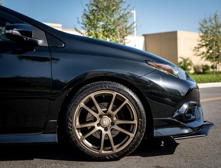 Close-up of a black car with an aftermarket bronze wheel, showing the front driver side and bumper detail.