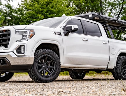 A white pickup truck with large, black off-road tires parked on a gravel surface, featuring a roof rack and surrounded by green trees.
