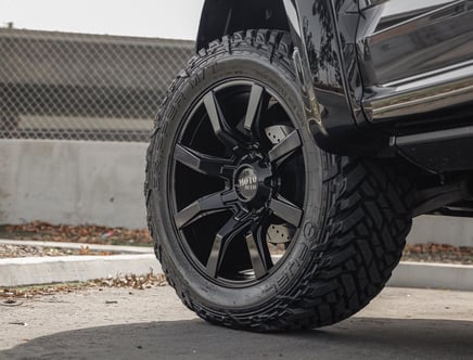 Close-up of a black off-road tire and wheel on a vehicle, with a visible "MOTO METAL" logo on the wheel hub, on a paved surface near a fence and curb.