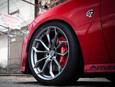 Close-up of the front wheel of a red car with a distinctive emblem on the fender, featuring large silver alloy rims and red brake calipers, parked in front of a chain-link fence with a "No Trespassing" sign.