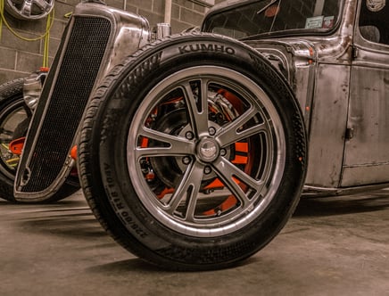 Close-up view of a shiny alloy wheel attached to a vintage car with a weathered, metallic body, featuring a Kumho tire in a garage setting.