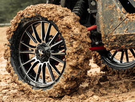 Close-up of a large off-road vehicle wheel covered in thick mud, highlighting the shiny black alloy rim and the rugged tire tread caked with dirt. The surrounding area is also muddy, indicating rough terrain.