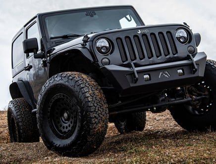 A black Jeep Wrangler with large off-road tires parked on uneven terrain, viewed from a low angle to emphasize its ruggedness.