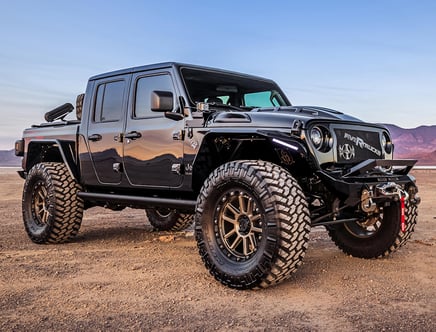 A black, customized off-road truck with large tires and a rugged grille is parked in a desert landscape with mountains in the background and a clear sky overhead.