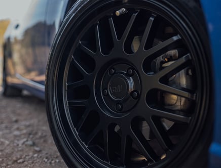 Close-up of a black alloy wheel with a multi-spoke design on a blue car, parked on a dirt road.