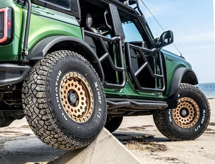 A green off-road vehicle with distinctive Gladiator X-Comp A/T tires and rugged suspension flexing on a concrete barrier near a beach.