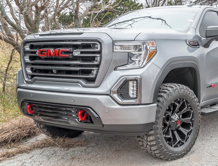 Front view of a silver GMC truck with a prominent red GMC logo on the grille, large off-road tires, and detailed headlight and bumper design.