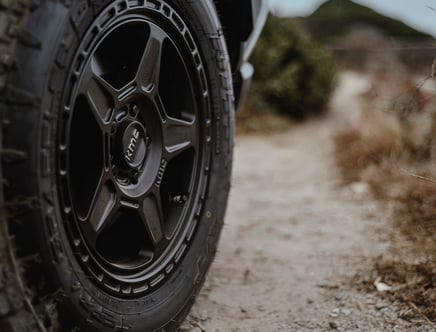 Close-up of a KMC black alloy wheel and tire on a gravel path with blurred natural surroundings.