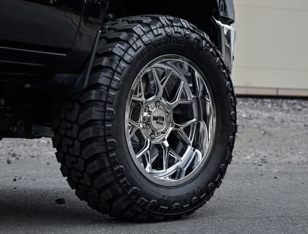 A close-up view of a large, rugged all-terrain tire on a black vehicle, featuring a shiny Moto Metal alloy wheel with a distinctive multi-spoke design.