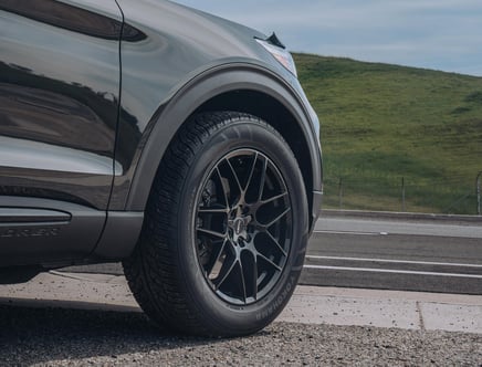 A close-up of a black car's front wheel with Yokohama tires and custom black alloy rims, parked on a roadside with a grassy hill in the background.