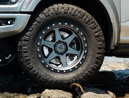 Close-up of an off-road vehicle's wheel with large, rugged tire treads, partially submerged in water and resting on rocks, showcasing the tire's branding and design details.
