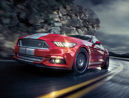 Red sports car with racing stripes driving on a wet, winding mountain road during a rainy evening.