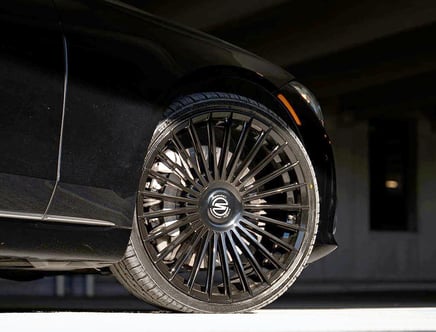 Close-up view of a stylish black car wheel with intricate multi-spoke rims, parked on a smooth surface under dim lighting.