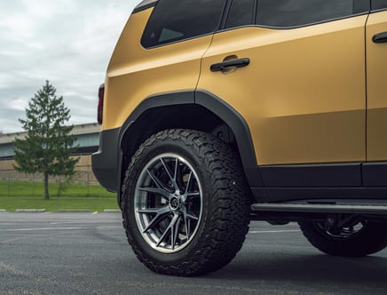Rear side view of a gold-colored SUV with large, rugged tires parked on a paved surface near a tree and a building in the background.