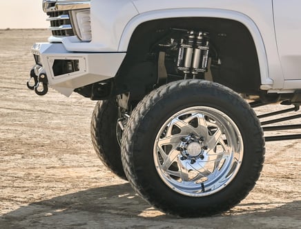 Close-up of a lifted pickup truck with large off-road tires and chrome wheels, featuring a custom suspension and front bumper with tow hooks, parked on a dirt surface.
