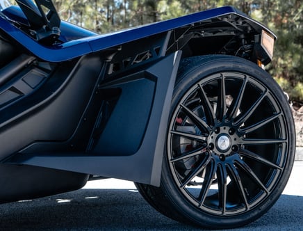 Close-up of a black alloy wheel with multiple thin spokes on a sleek vehicle, set against a blurred natural background.