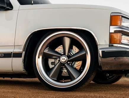 Close-up of the front wheel of a white car, featuring a large, metallic, five-spoke rim with a "U.S. Mags" logo at the center, parked on a dirt surface.