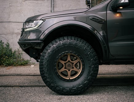 Close-up of a truck's front left wheel and fender, featuring a large off-road tire with bronze-colored rims, parked on a gravel surface next to a concrete wall with some greenery visible.