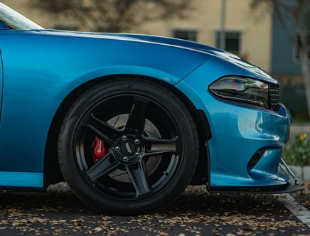 Close-up view of a blue car's front wheel and side, featuring a black alloy rim with a red brake caliper.