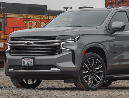 Close-up of the front and side view of a gray Chevrolet Tahoe SUV, with a sleek grille design, modern headlights, and visible alloy wheels, parked on gravel in front of a blurred orange and black background possibly showing a train or industrial setting.