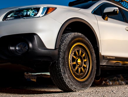 Angled view of a silver Subaru Outback with aftermarket golden wheels on a gravel path.