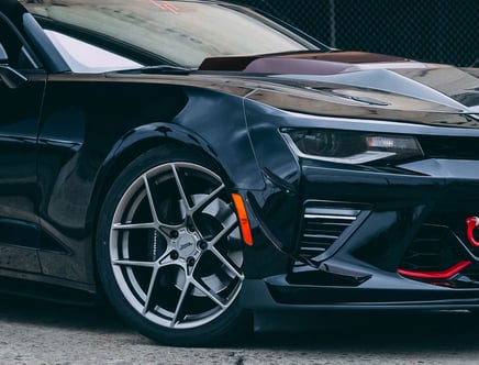 Close-up of the front side of a black sports car with silver alloy wheels, aggressive styling, and a red tow hook.
